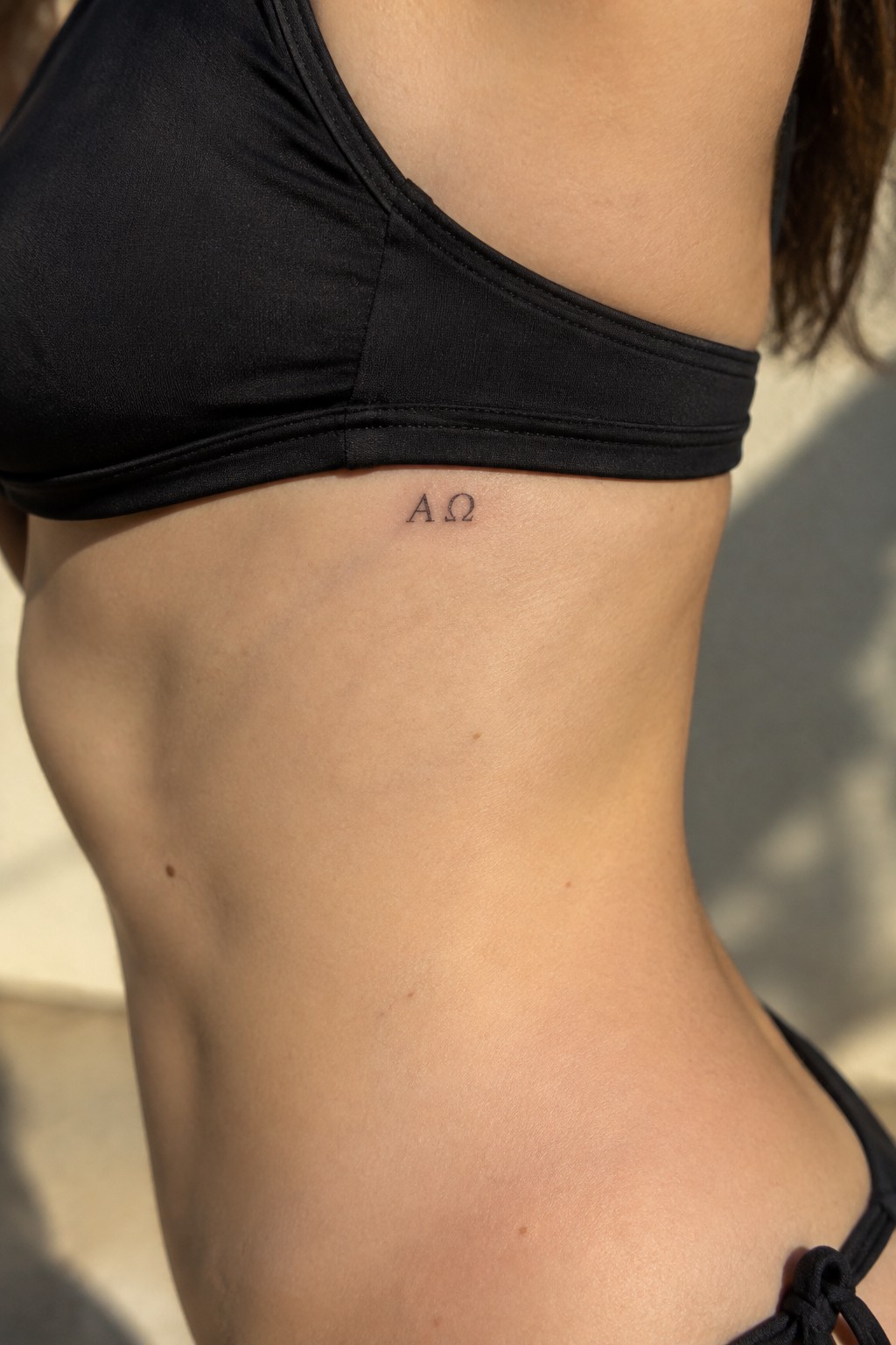 Woman's torso in black bikini with A Ω tattoo below ribs in sunlight