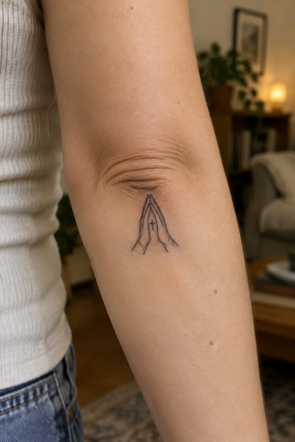 Praying hands tattoo with cross on elbow, white ribbed top and blue jeans.