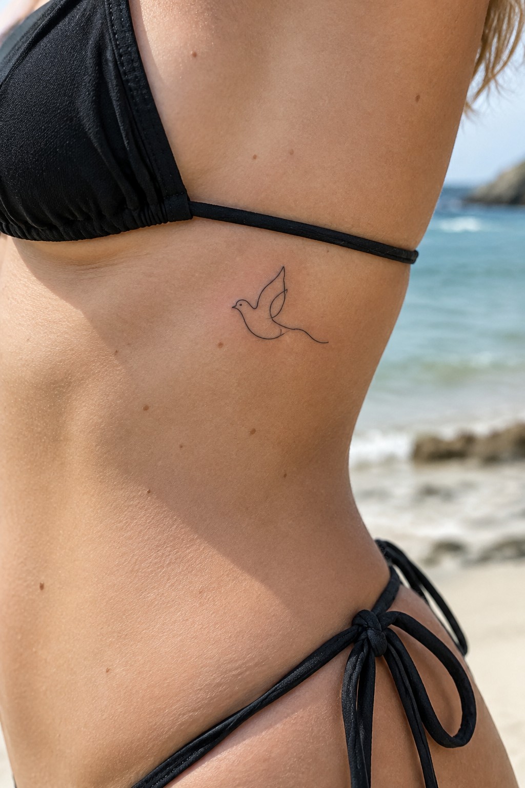 Minimalist single line dove tattoo on a woman's ribcage area in a black bikini at the beach.