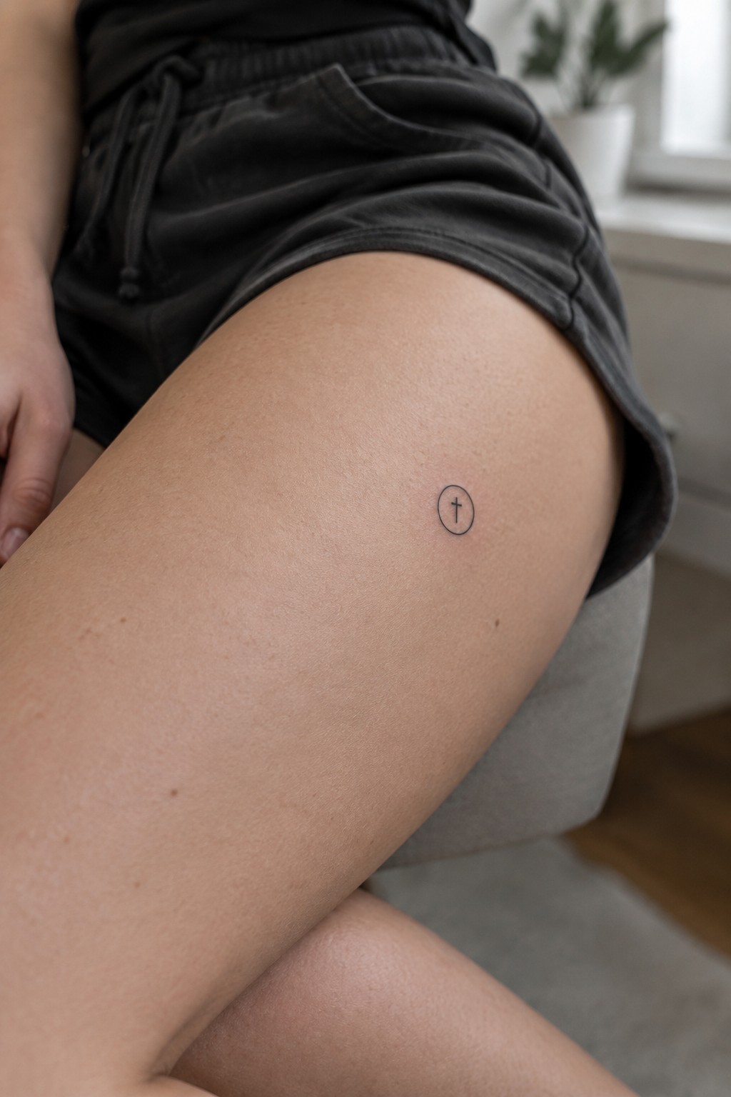 Small oval cross tattoo on thigh in black shorts on gray chair