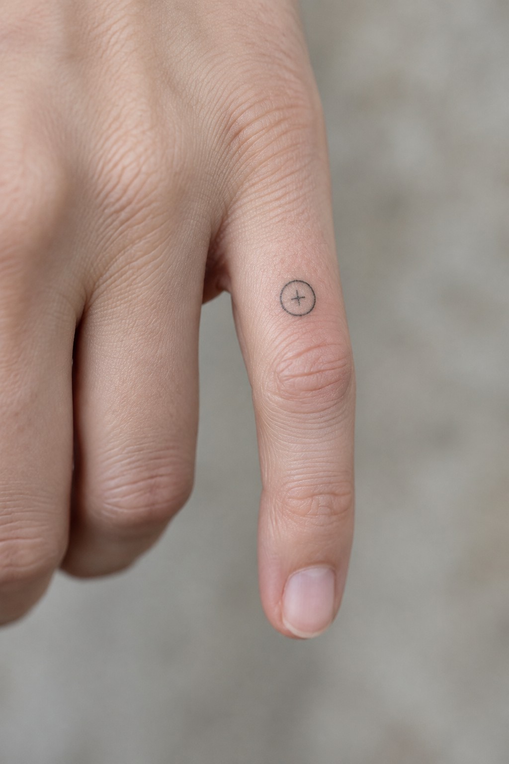 Close-up of a finger with a small black plus tattoo inside a circle.