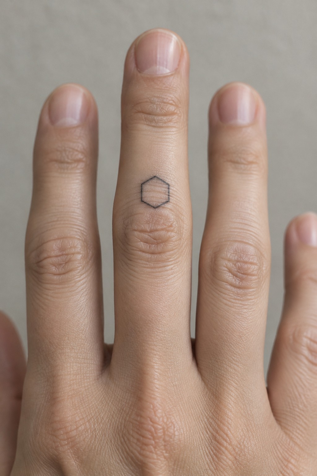 Fingers of a hand with a small black hexagon tattoo on the middle finger.