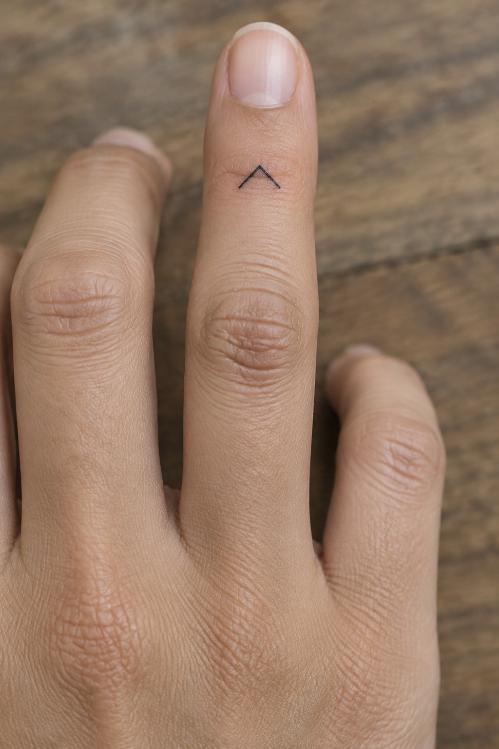 Hand on wooden surface with small black chevron tattoo on middle finger.