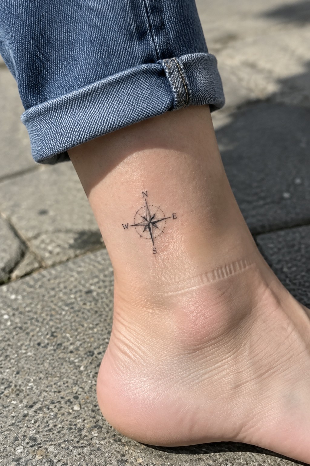 Small black compass tattoo with N, S, E, and W markings on the inner ankle above the heel.