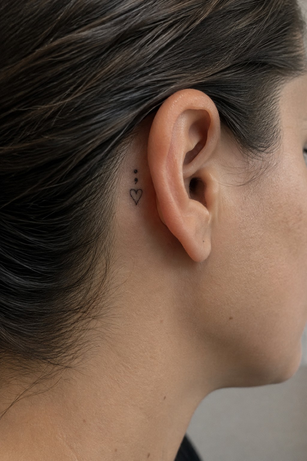 Small black fine-line heart tattoo with two dots above it behind a woman’s ear.