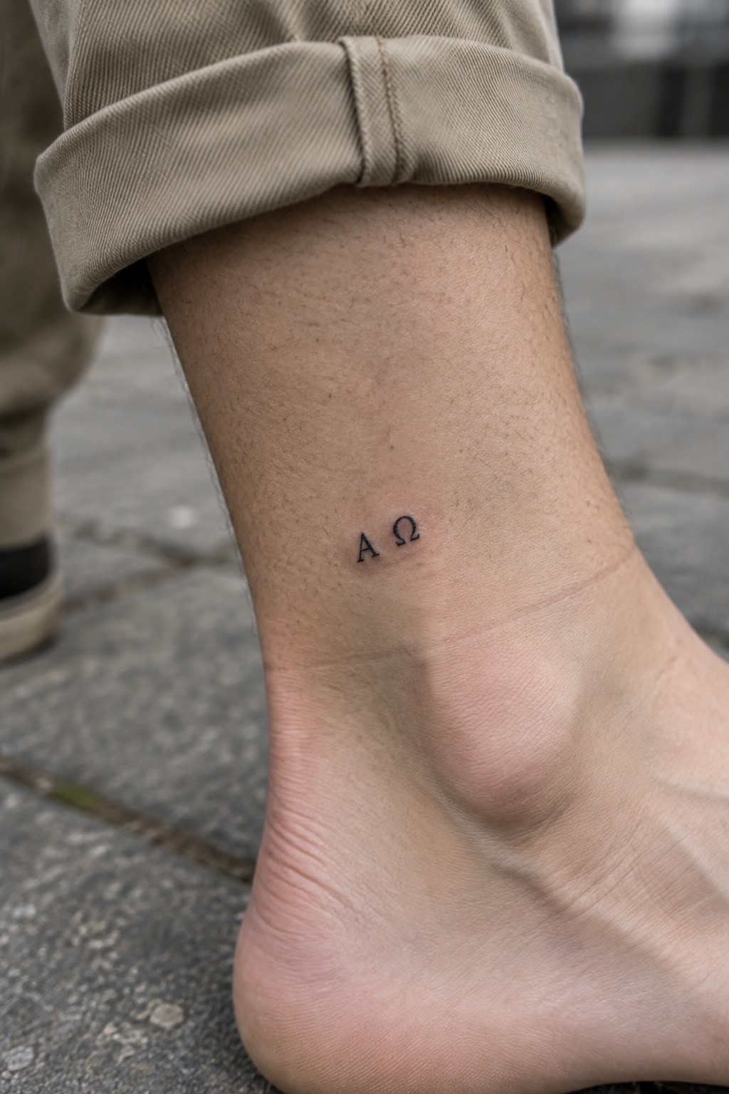 Minimalist black ink alpha and omega tattoo on the inner ankle.