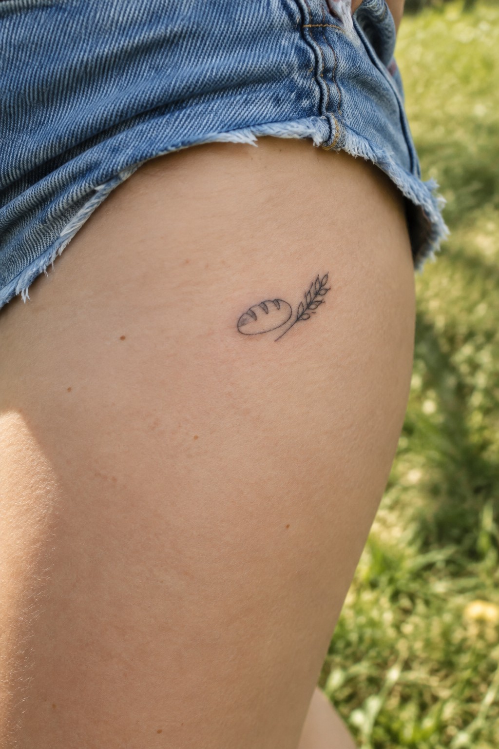 Frayed blue denim shorts and small bread-wheat tattoo on thigh against blurred grass