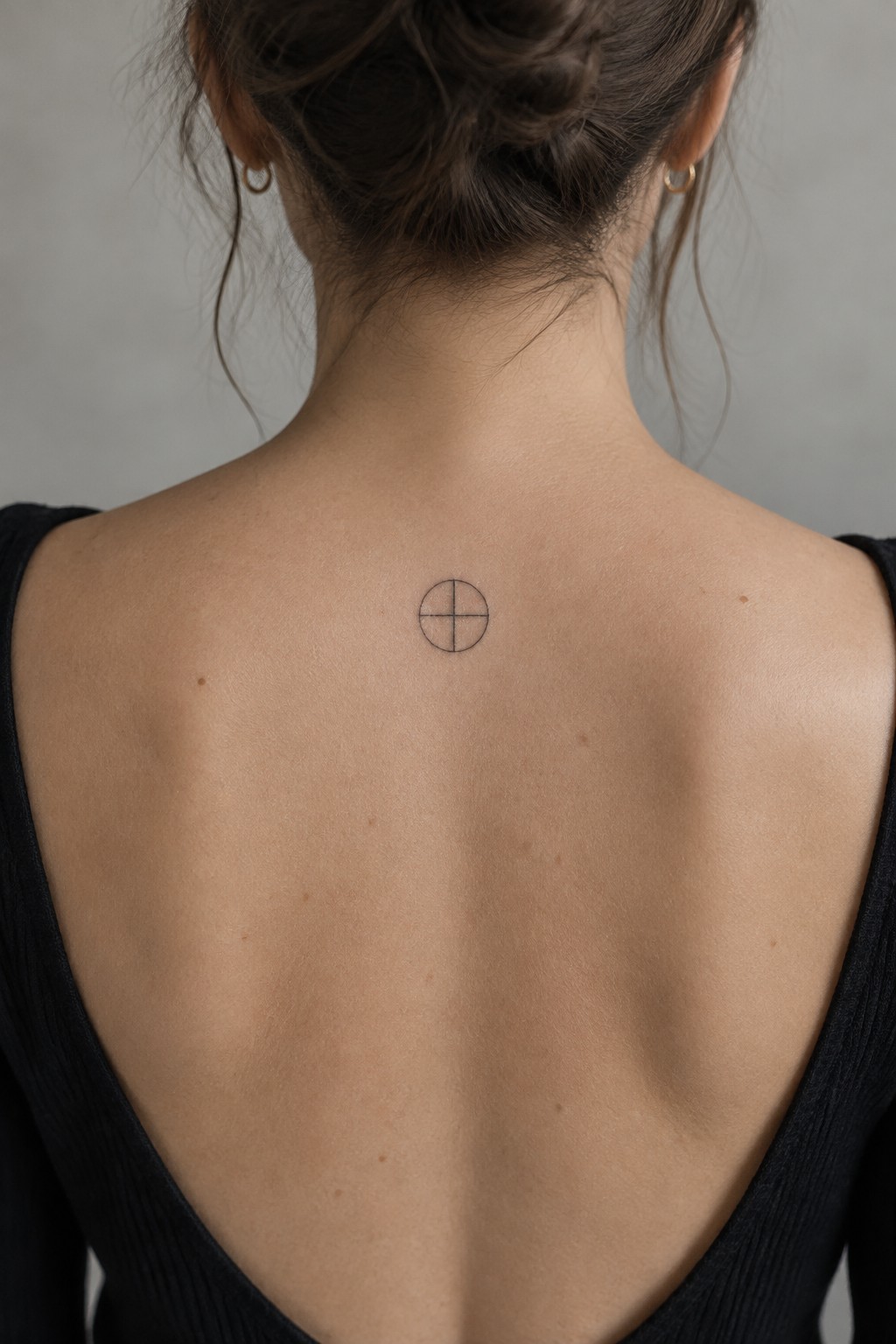 Woman's back with small cross-in-circle tattoo, brown bun, black low-back top