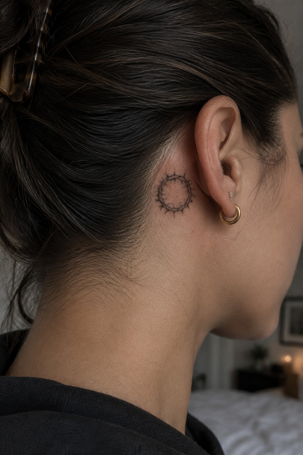 Close up of a small circular tattoo with thin spikes behind a woman's ear, done in minimalist black linework.