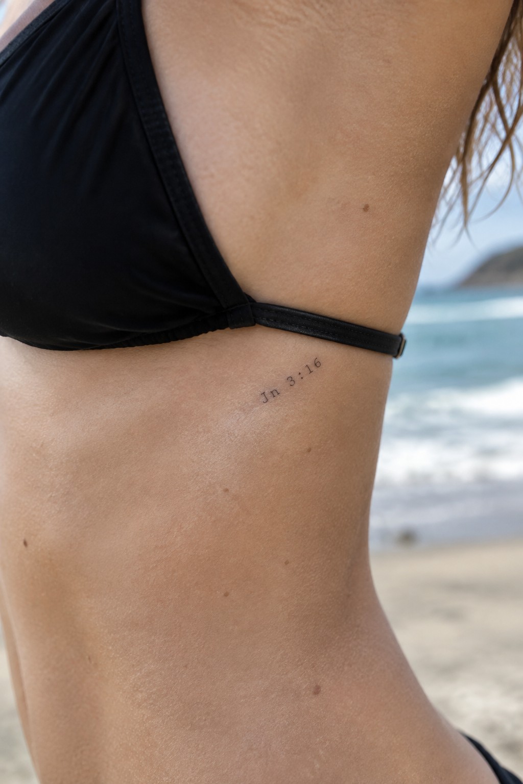 Small black text tattoo of Jn 3:16 on the side torso near the ribs on a person in a black swimsuit at the beach