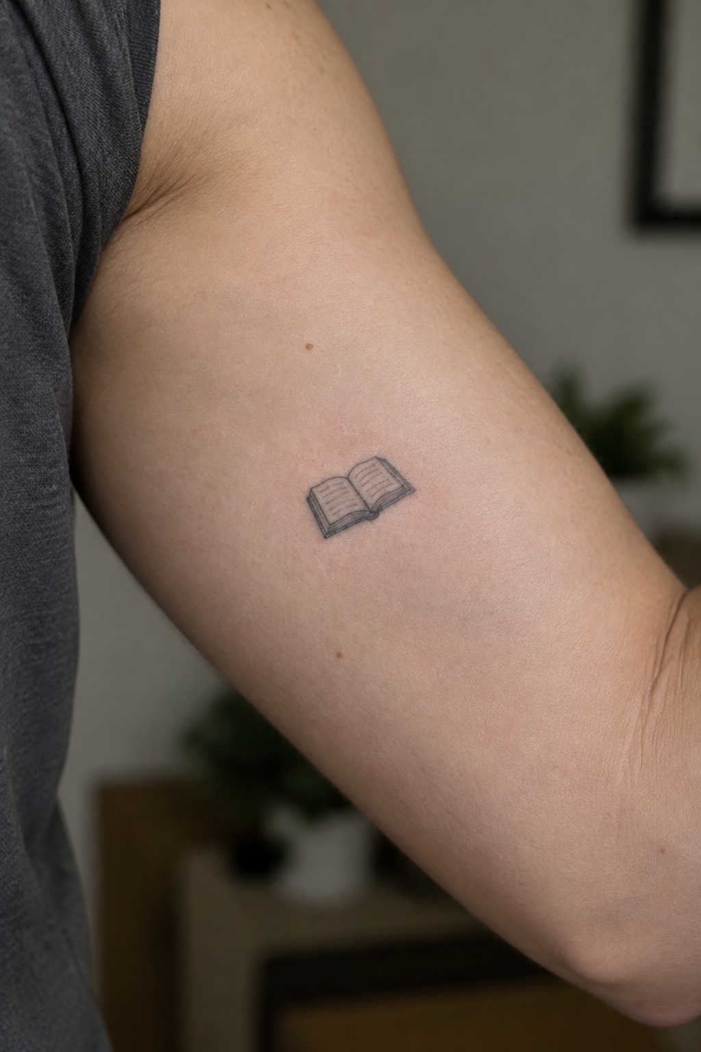 Arm in gray shirt with small open book tattoo on upper arm