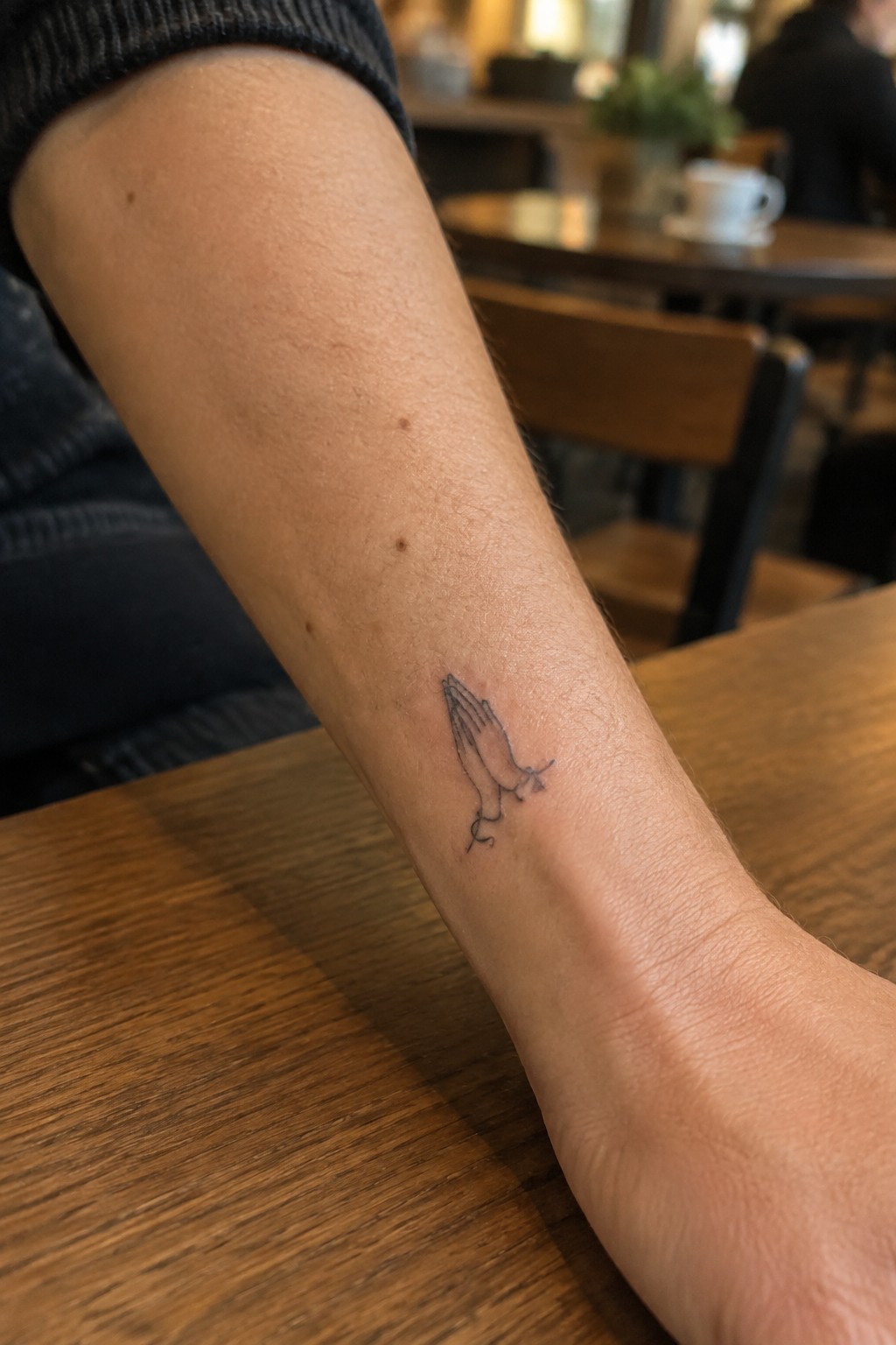 Arm with small praying hands tattoo resting on wooden cafe table