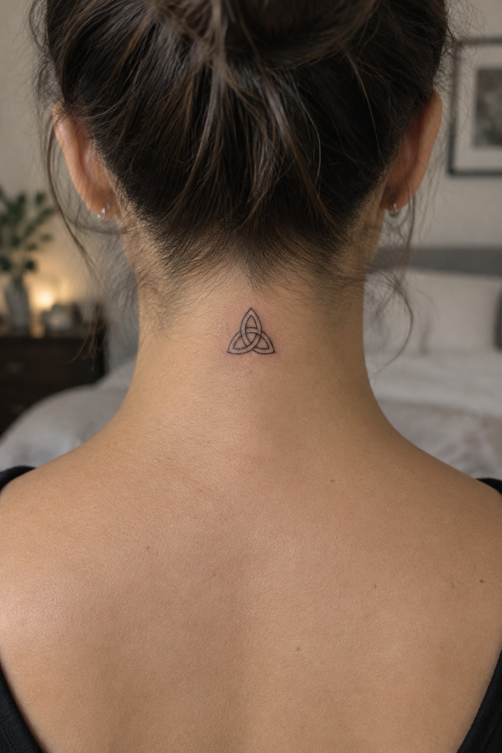 Woman's neck with triquetra tattoo below brown hair bun, black top, soft bedroom background