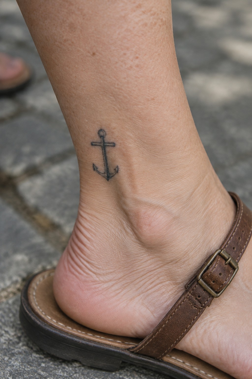Black anchor tattoo on ankle above brown leather sandal buckle