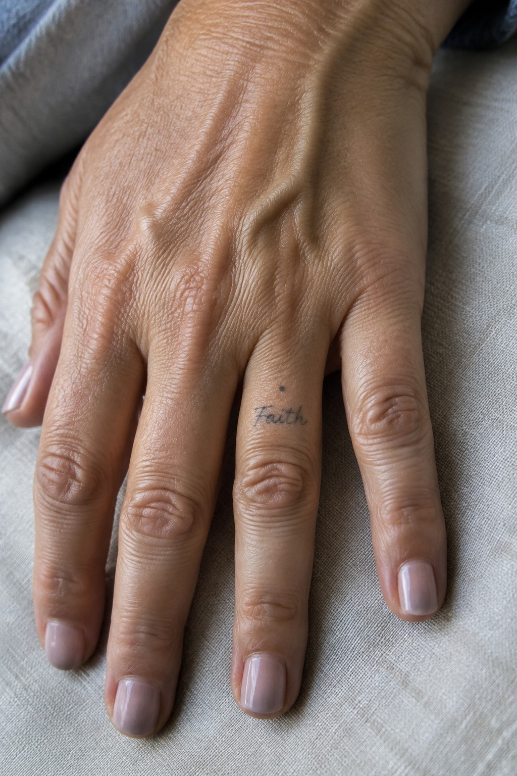Hand with cursive Faith tattoo on ring finger, pink nails on light fabric