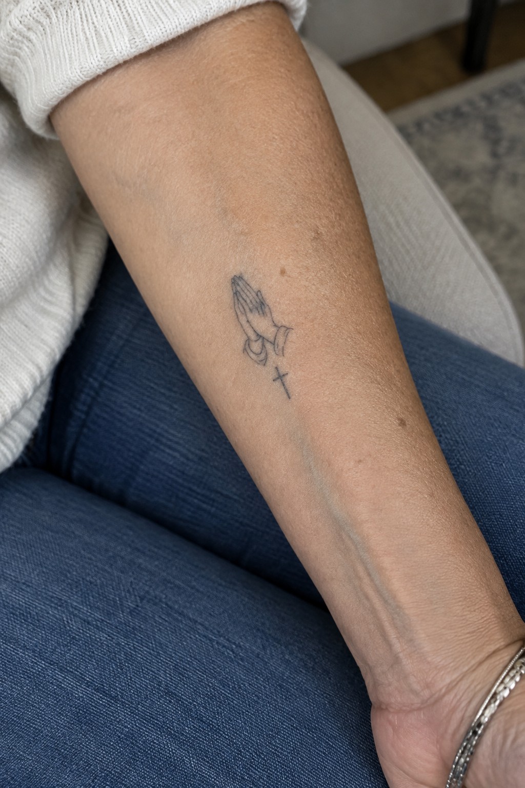 Person's forearm with praying hands and cross tattoo, white sweater, blue pants