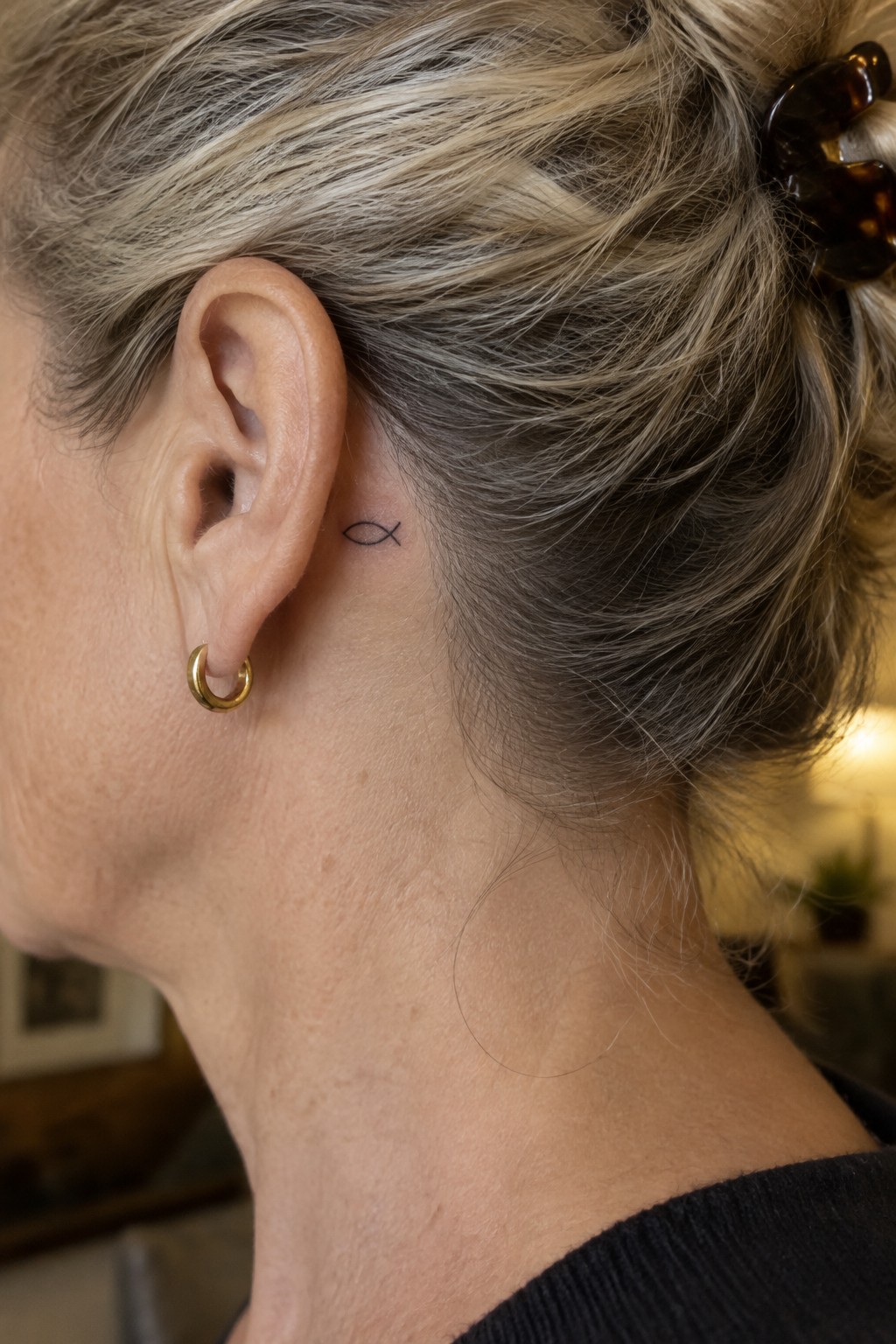 Woman with blonde-gray bun, fish tattoo behind ear, gold hoop earring, black top