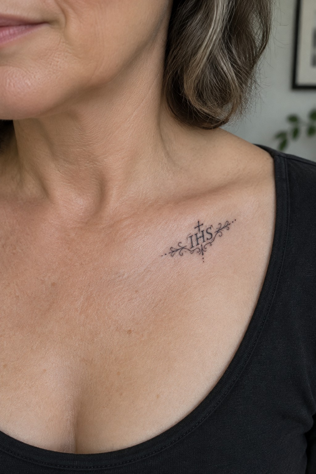 Ornate black IHS tattoo with cross and flourishes on collarbone above black top