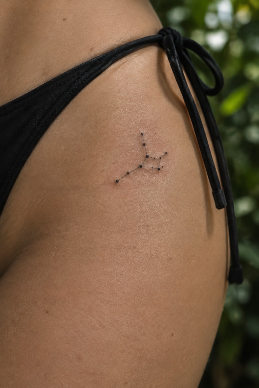 Small black fine-line constellation tattoo on the upper hip beside a black bikini tie.