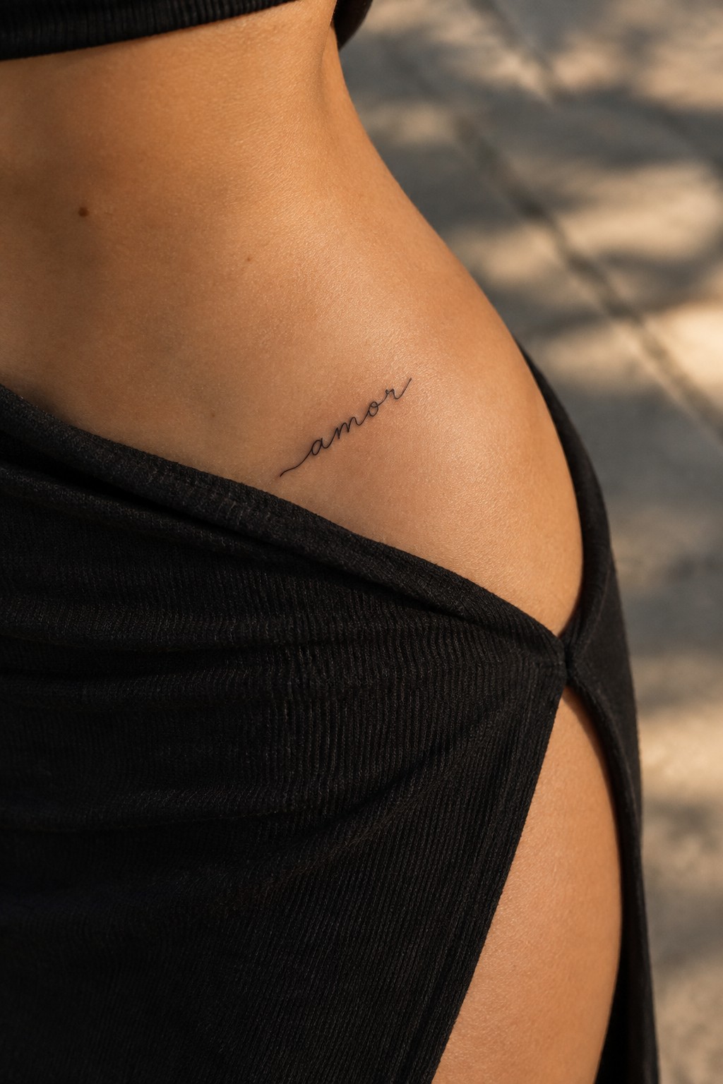 Small fine-line cursive “amor” tattoo above the hip, partly framed by black clothing.