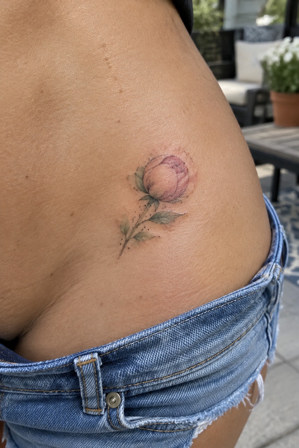 Small pink rosebud tattoo with a fine stem and muted green leaves on the side of a woman’s hip above denim shorts.