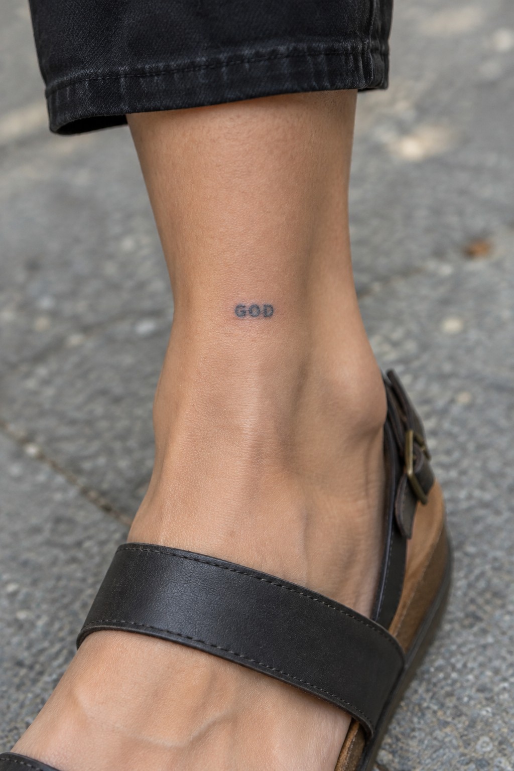 Ankle with small black GOD tattoo above black leather sandal strap on pavement