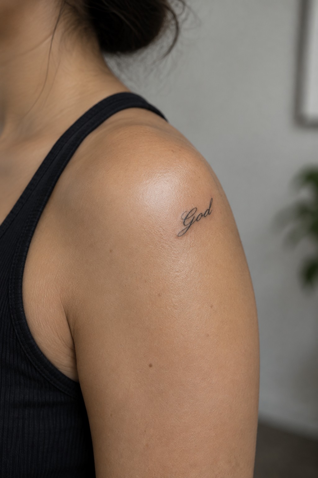 Small cursive tattoo of the word God on the shoulder of a person wearing a black tank top.