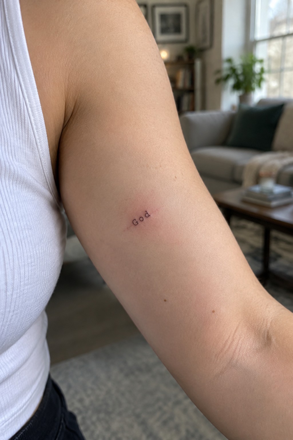 Small fine line tattoo of the word God in delicate script on the upper arm