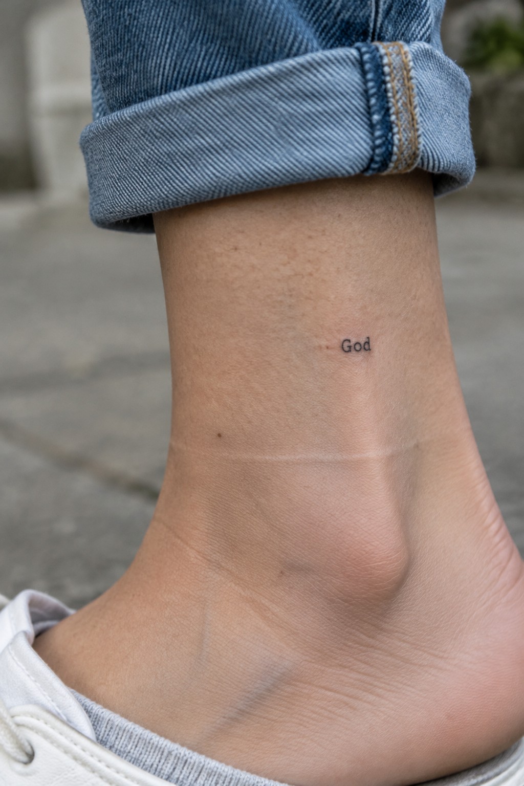 Close-up of ankle with 'God' tattoo below rolled blue jeans cuff and white shoe.