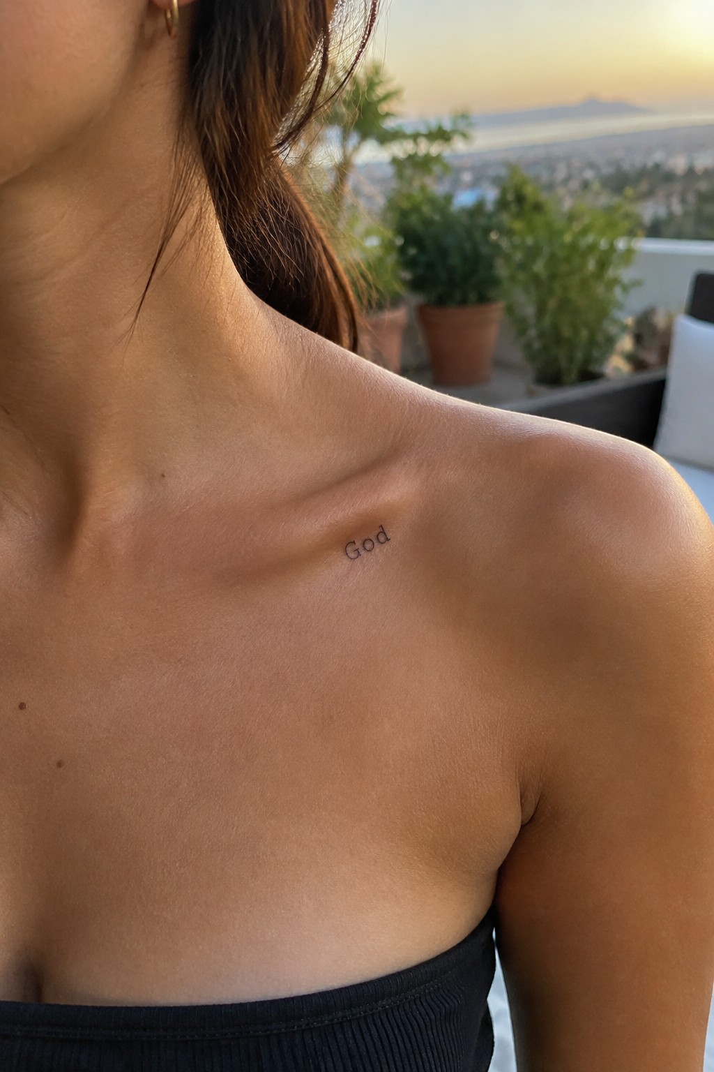 Close up of a small single word tattoo reading God placed on the collarbone in minimalist lettering.