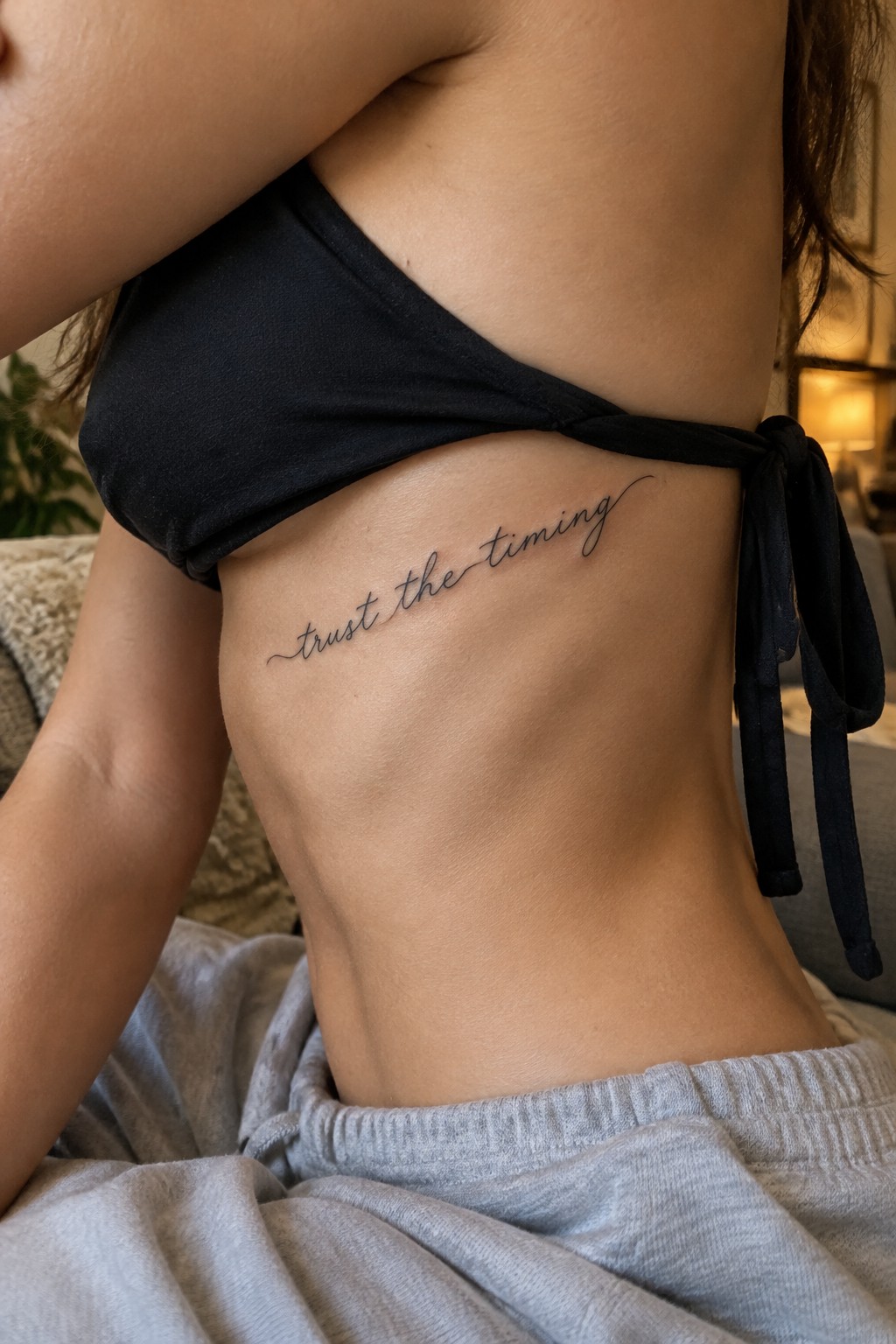 Fine-line cursive tattoo reading “trust the timing” along the side of the ribs.