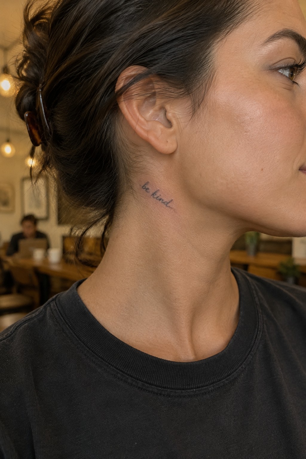 Small fine-line “be kind” script tattoo on the side of a woman’s neck below her ear.