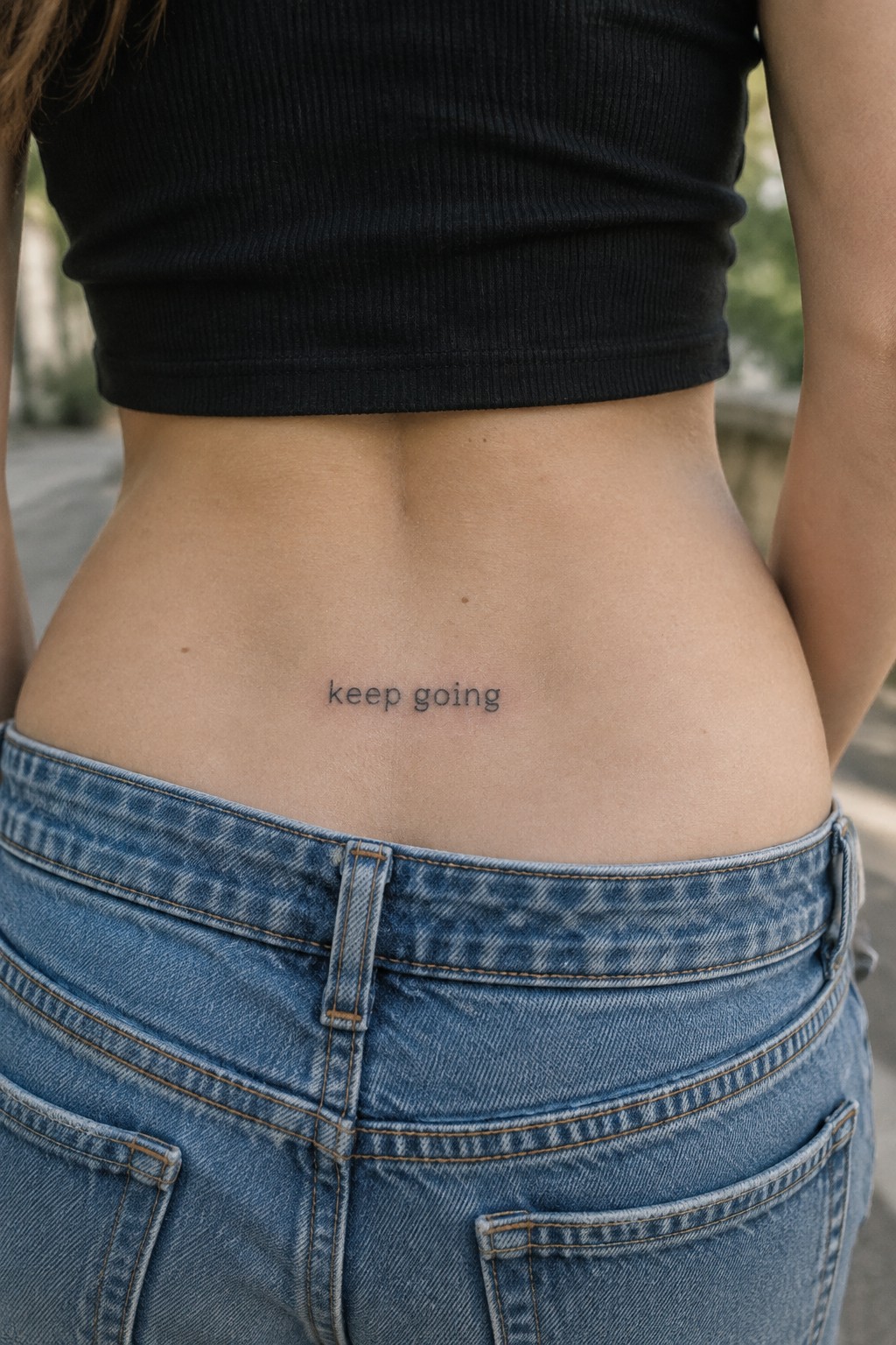 Small lowercase “keep going” script tattoo centered on a woman’s lower back above blue jeans.