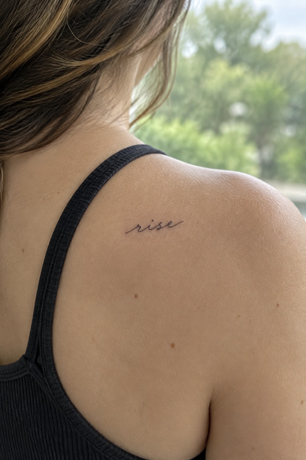 Small fine-line cursive “rise” tattoo on a woman’s shoulder.