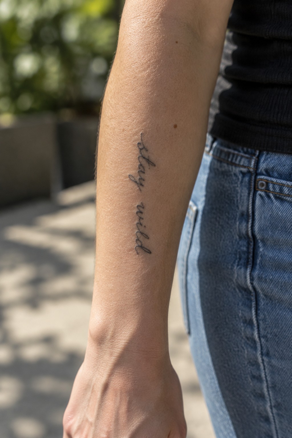 Fine-line cursive “stay wild” tattoo running vertically along the inner forearm.