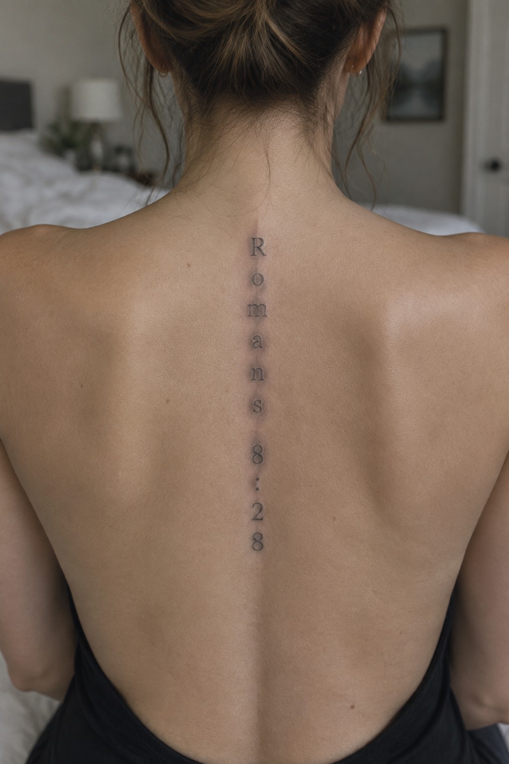 Woman's bare back with vertical Romans 8:28 tattoo along spine in bedroom