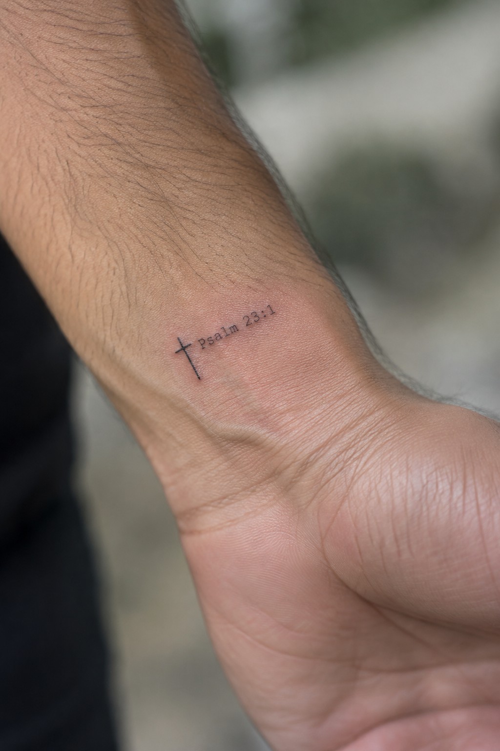 Small black cross tattoo with Psalm 23:1 text on the inner wrist