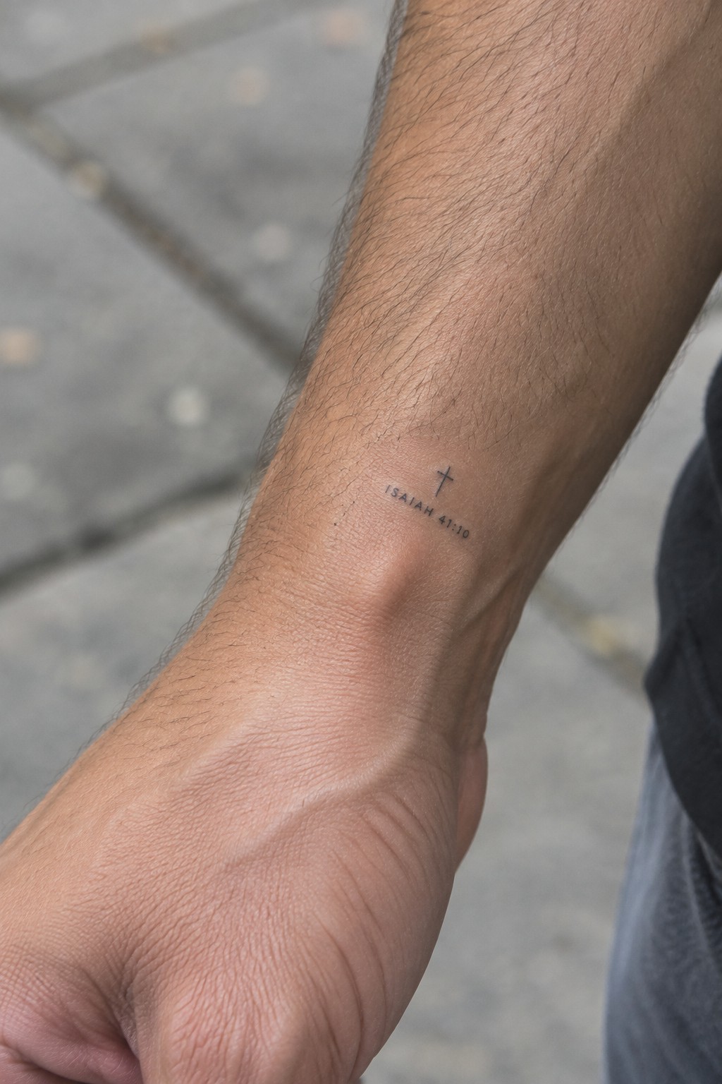Hairy wrist with small cross tattoo and Isaiah 41:10 text above clenched fist