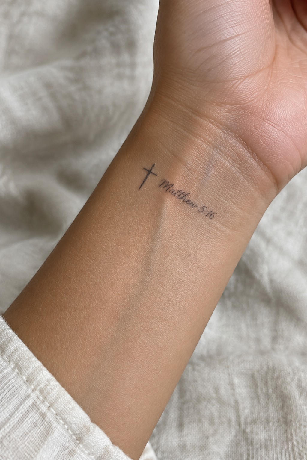 Small black cross tattoo beside the text Matthew 5:16 on the inner wrist of a light skin tone arm resting on white fabric