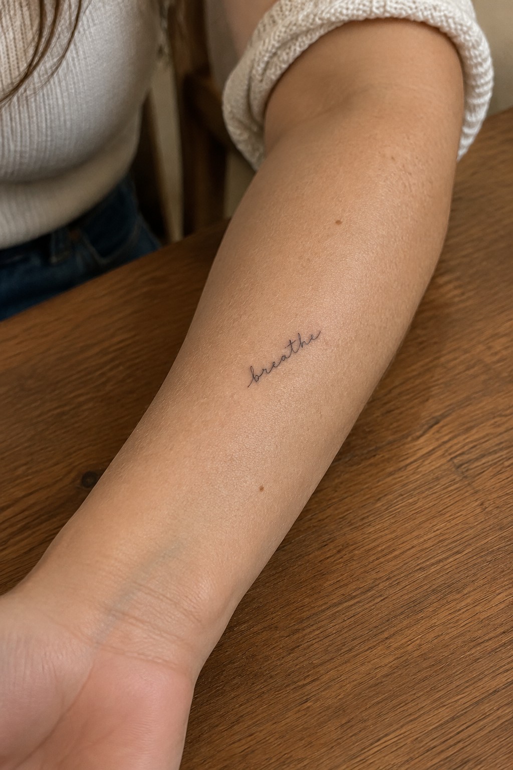 Small fine-line “breathe” script tattoo on the inner forearm.