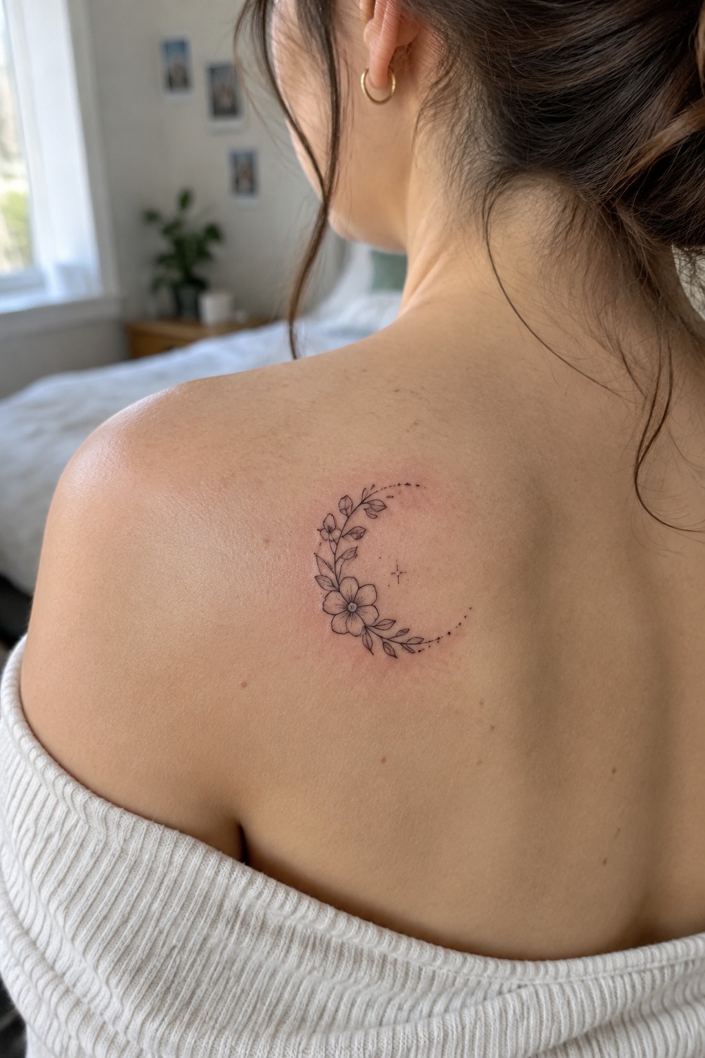 Fine-line crescent moon tattoo with a flower, leaves, and dotted details on a woman’s upper back near the shoulder.