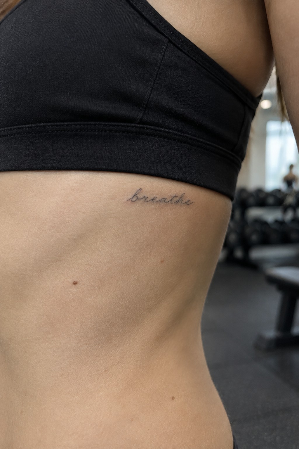 Small fine-line cursive “breathe” tattoo beneath a black sports bra on the ribcage.