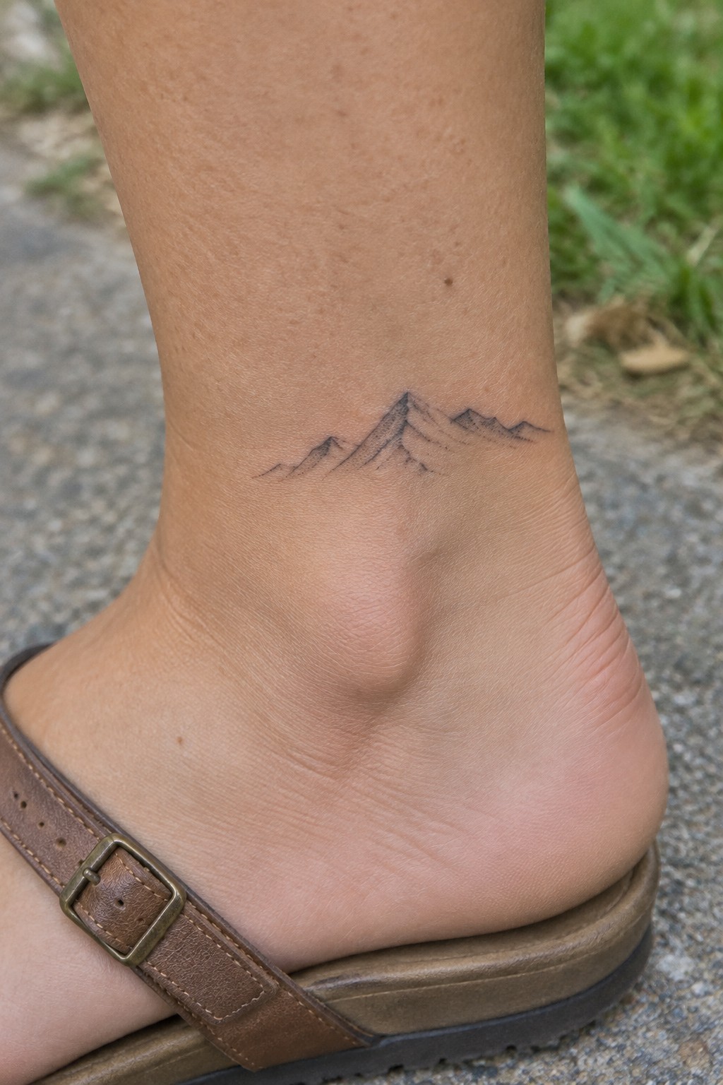 Small fine-line mountain range tattoo above the ankle, with lightly shaded peaks.
