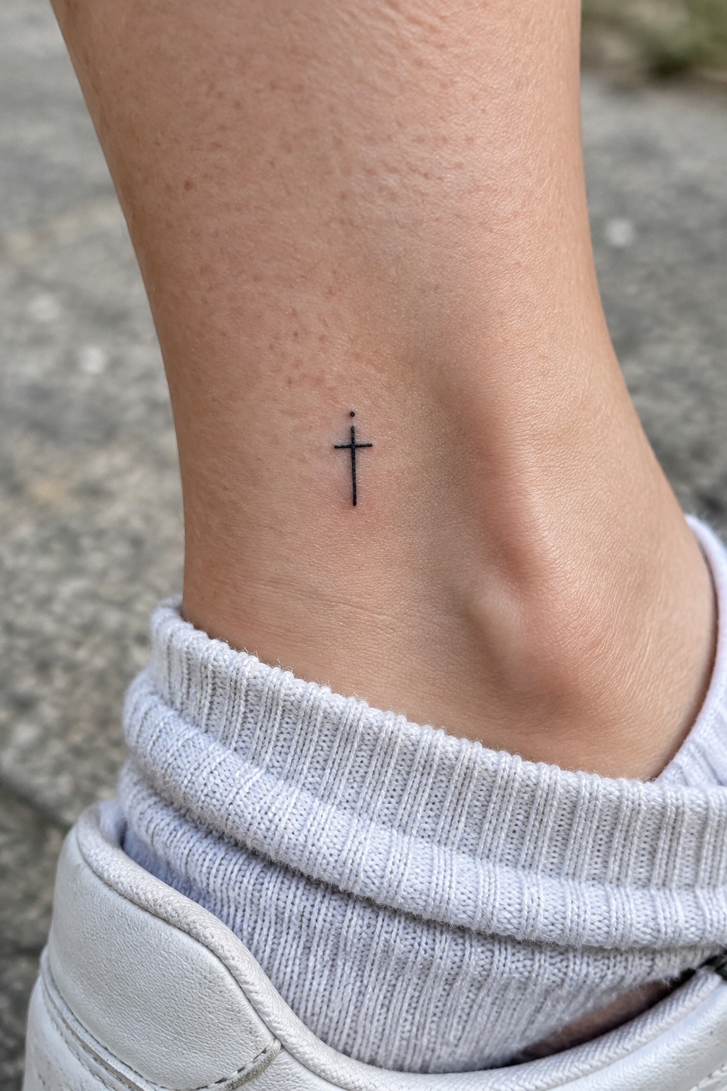 Small black cross tattoo on ankle above white ribbed sock and shoe.