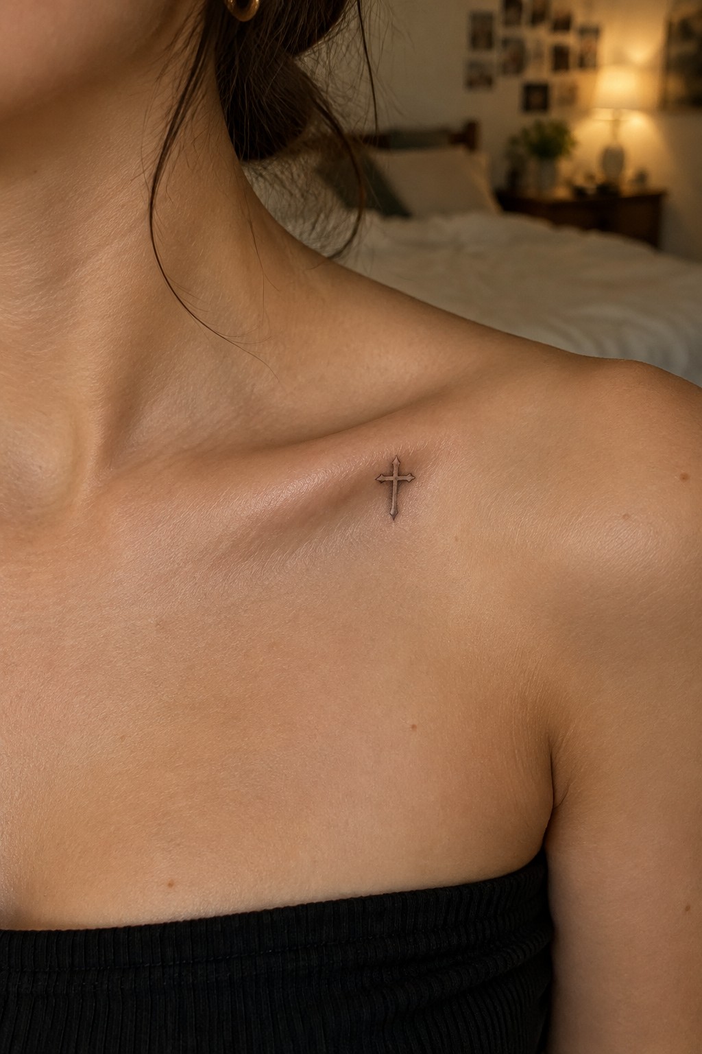 Small cross tattoo on collarbone above black top in bedroom setting.