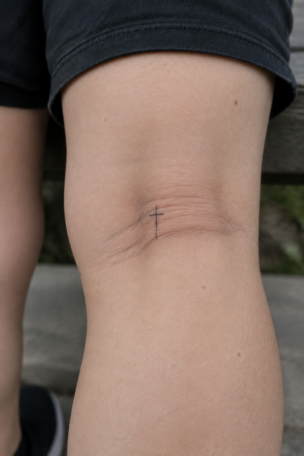 Small black cross tattoo on the knee in minimalist line style