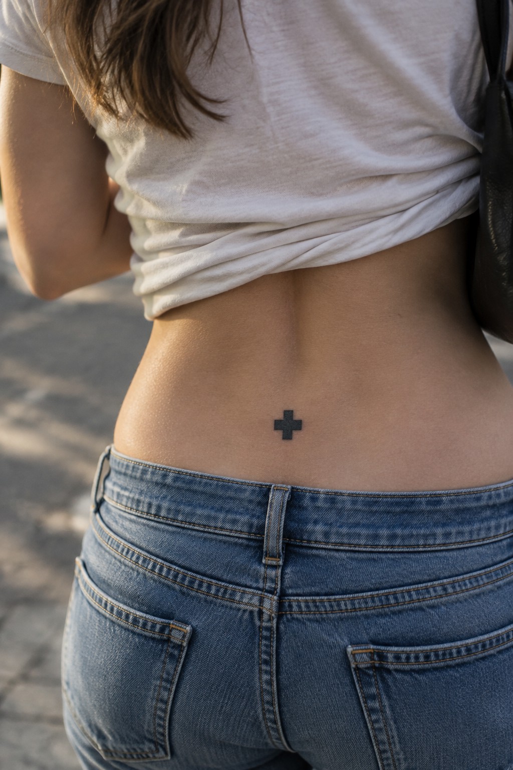 Woman's lower back with small black cross tattoo above blue jeans, white shirt lifted.