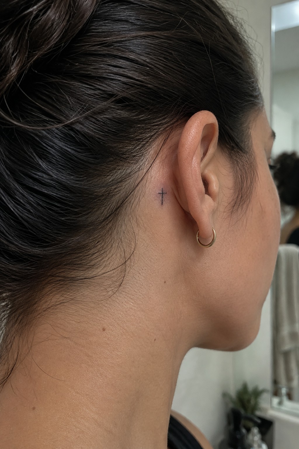 Small black cross tattoo behind the ear on dark hair pulled into a bun