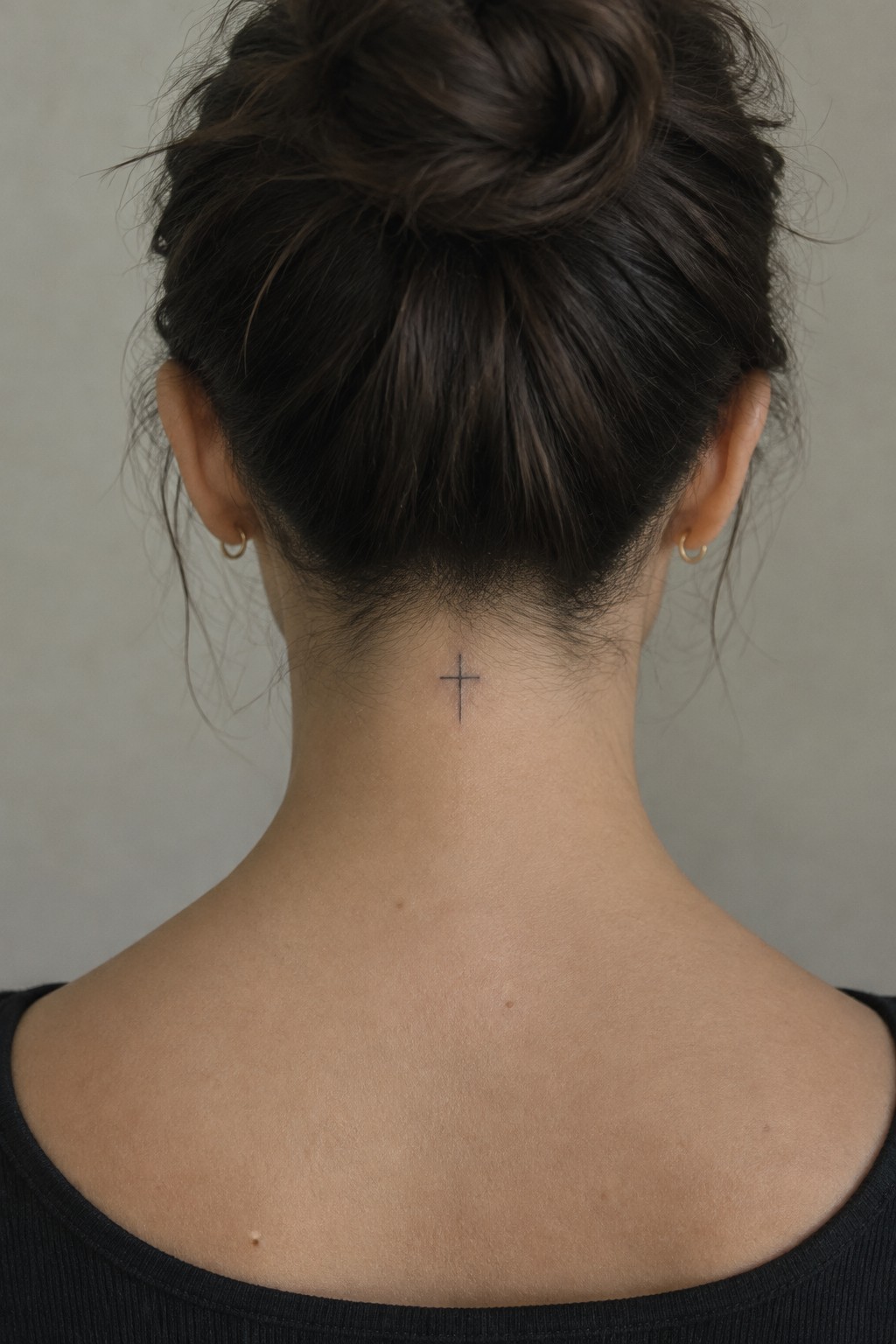 Woman's dark hair in bun above small cross tattoo on neck, black top