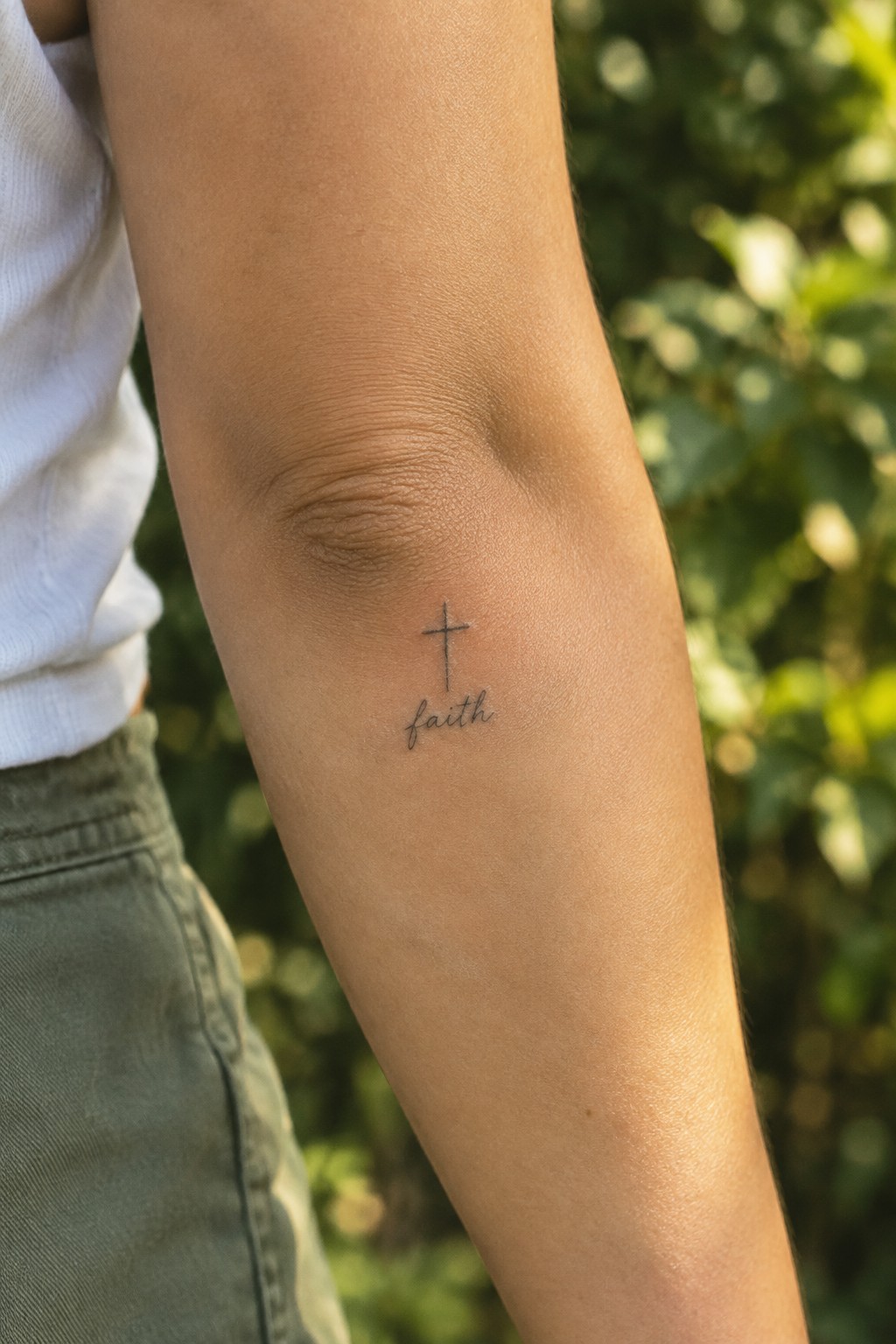Small cross and faith tattoo on bare arm with white shirt and green pants outdoors
